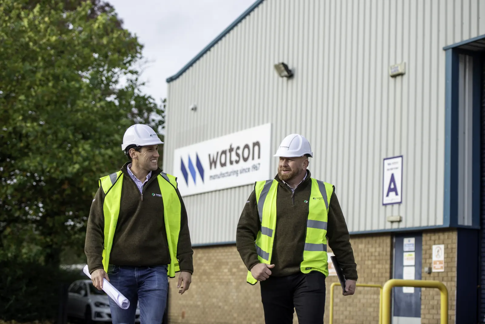 Two Cornerstone managers in hard hats and hi-vis walking past the Watson factory signage