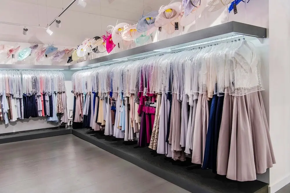 Boutique display rails of occasion dresses beneath a lit shelf of hats