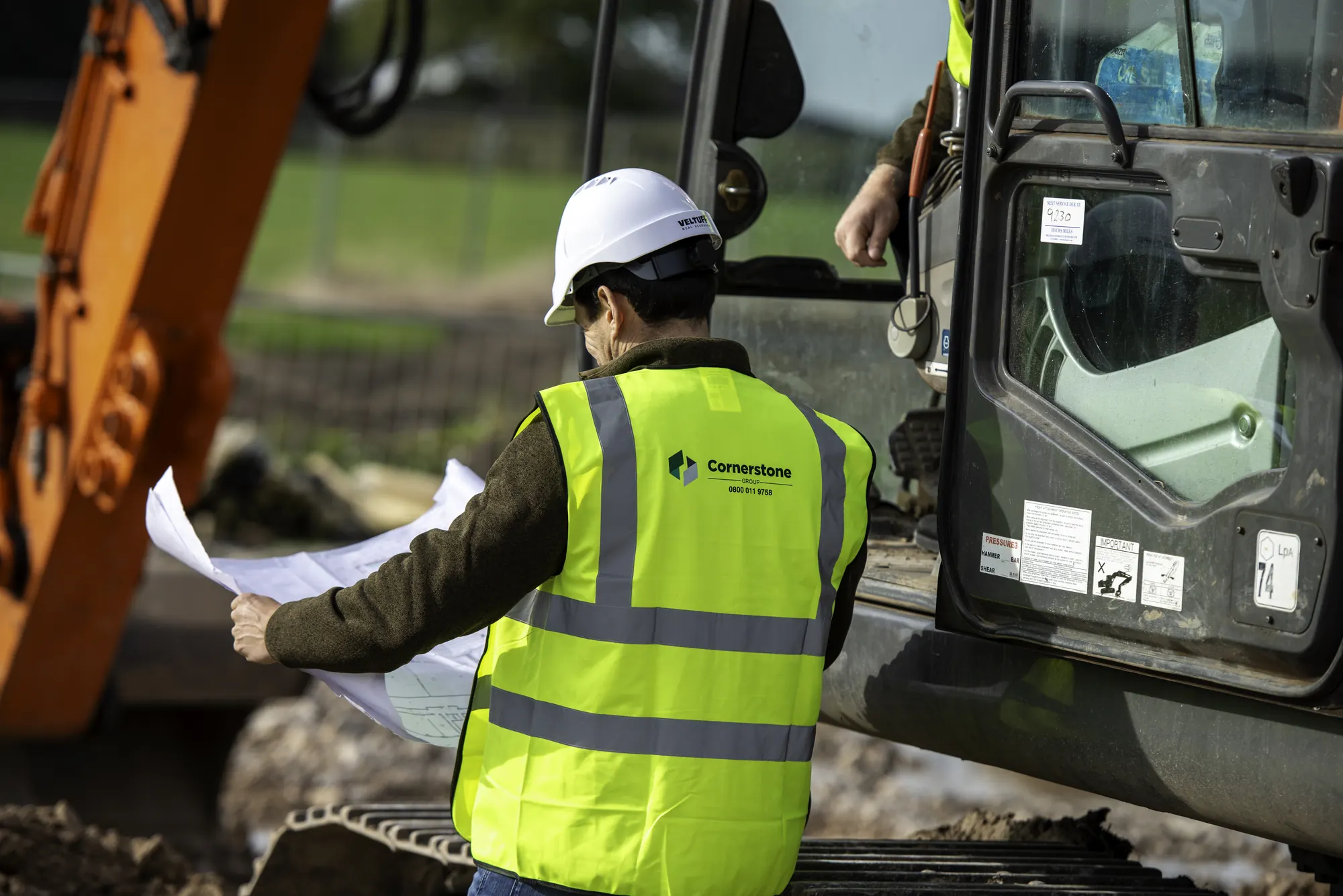 Cornerstone site manager in a hard hat and branded hi-vis checking plans beside an excavator