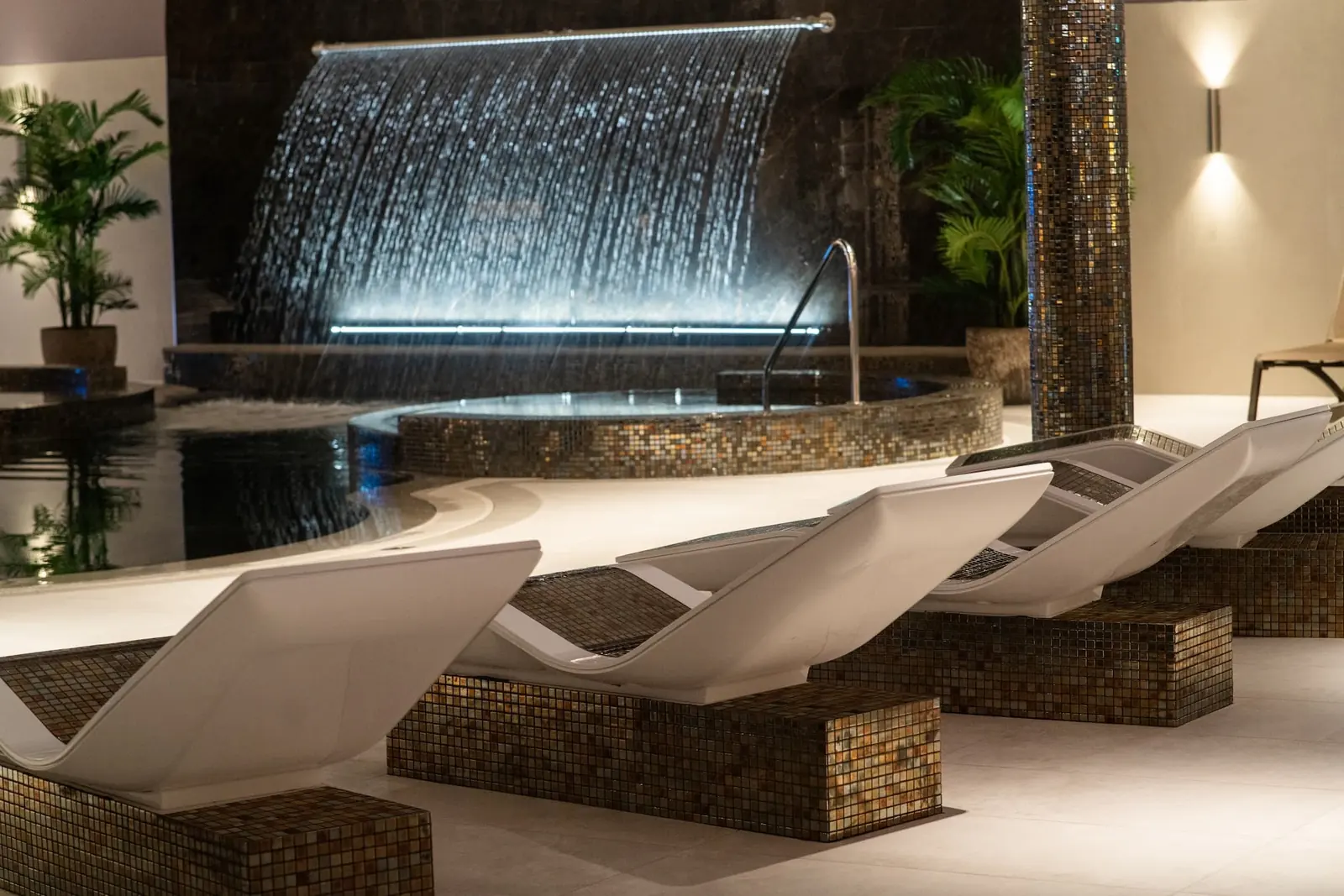 Sculpted white loungers on mosaic plinths beside a lit cascading water feature in Crow Wood spa