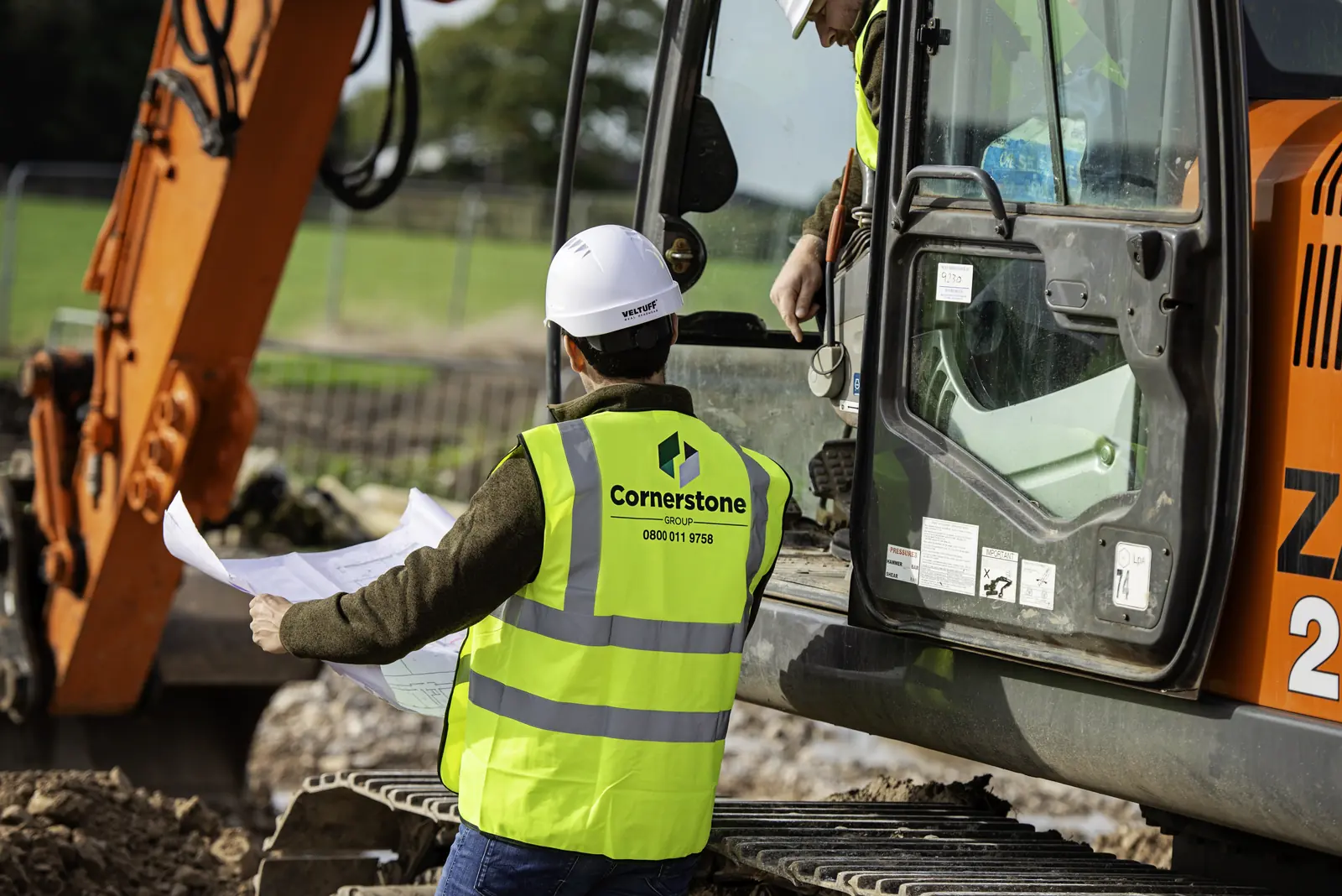 Cornerstone site manager in hard hat and branded hi-vis reading plans beside a tracked excavator