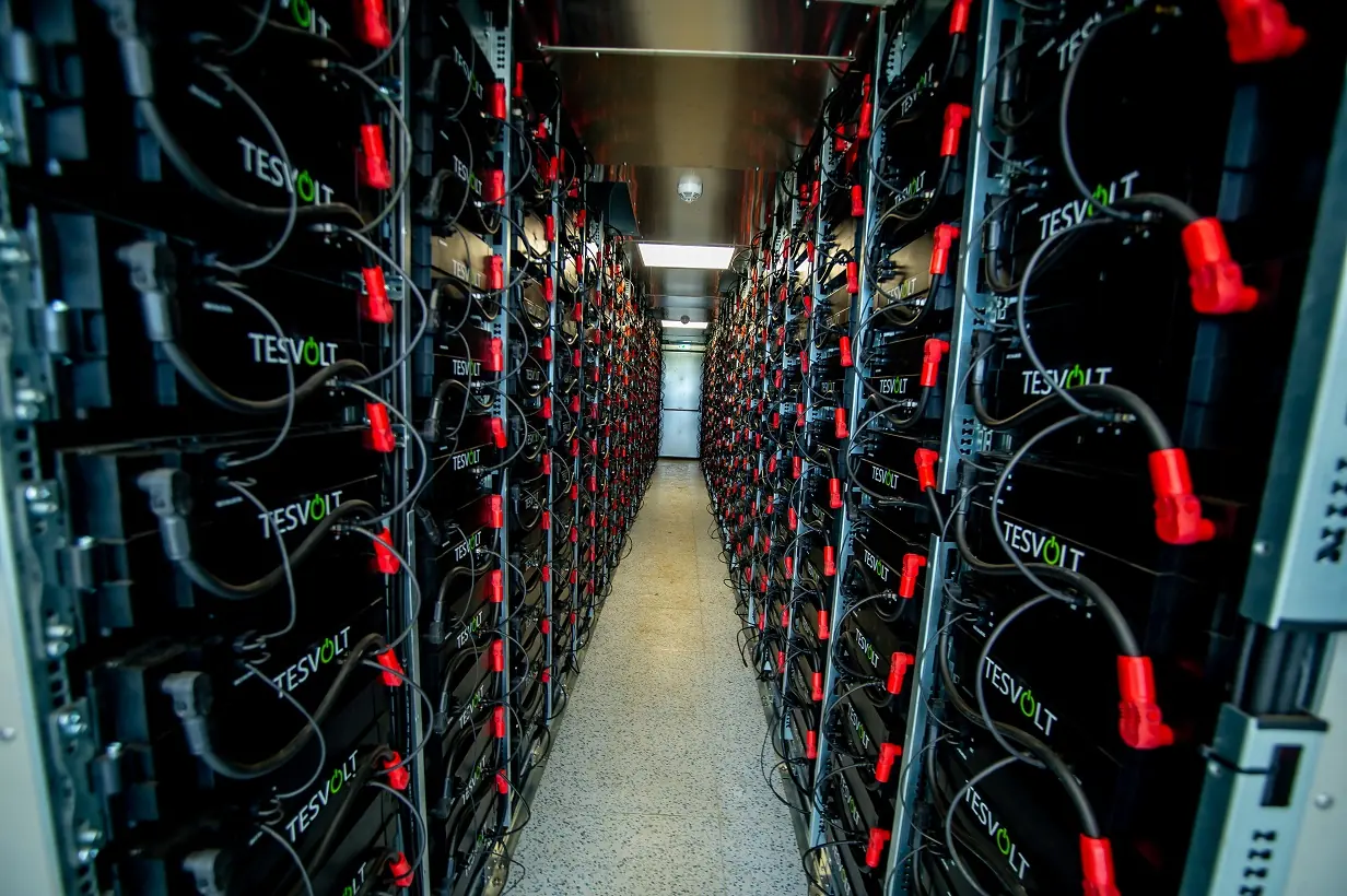 Rows of battery storage modules with red connectors in a narrow plant room aisle